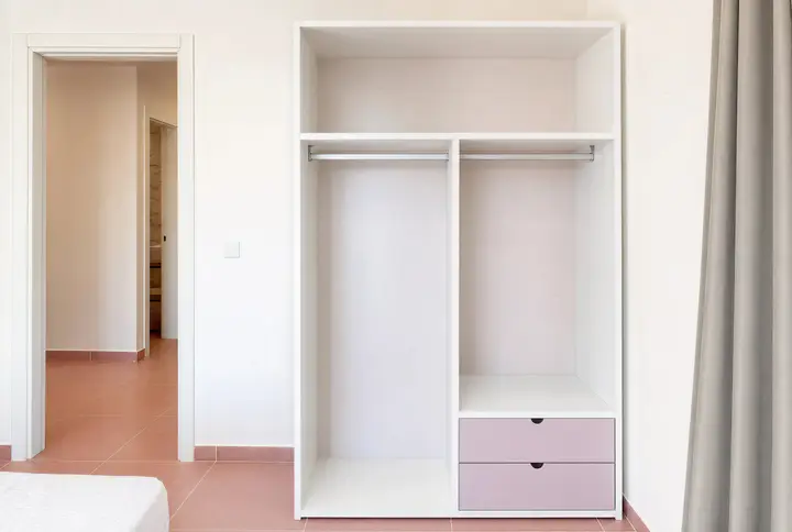 Open white wardrobe with hanging rails and pink drawers beside a doorway